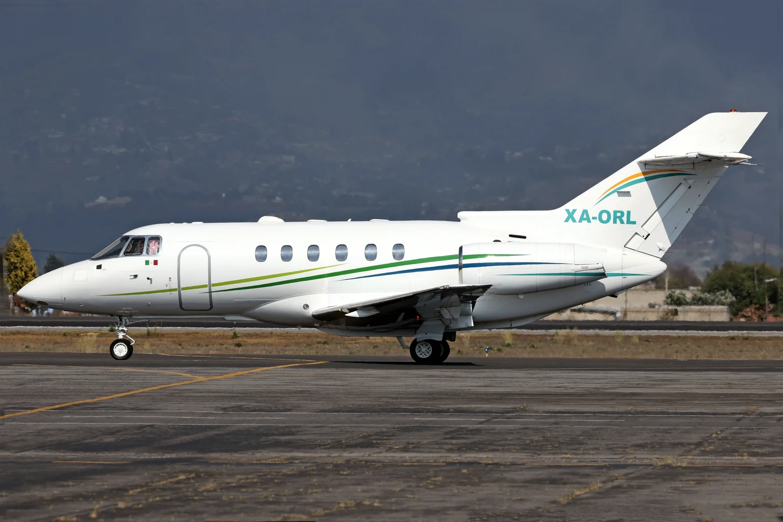 A white Hawker 800XP with green and blue stripes taxiing side-on at Toluca, Mexico, under a dark storm sky.