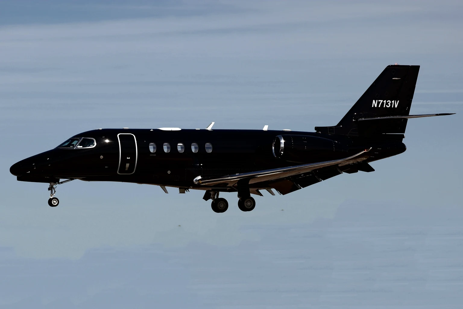 A gloss-black Cessna Citation Latitude on approach with its gear down against a pale blue sky