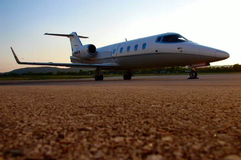 A white Learjet 55 parked on the apron at dusk, shot low from the tarmac with the sky turning pink behind the winglet and T-tail.