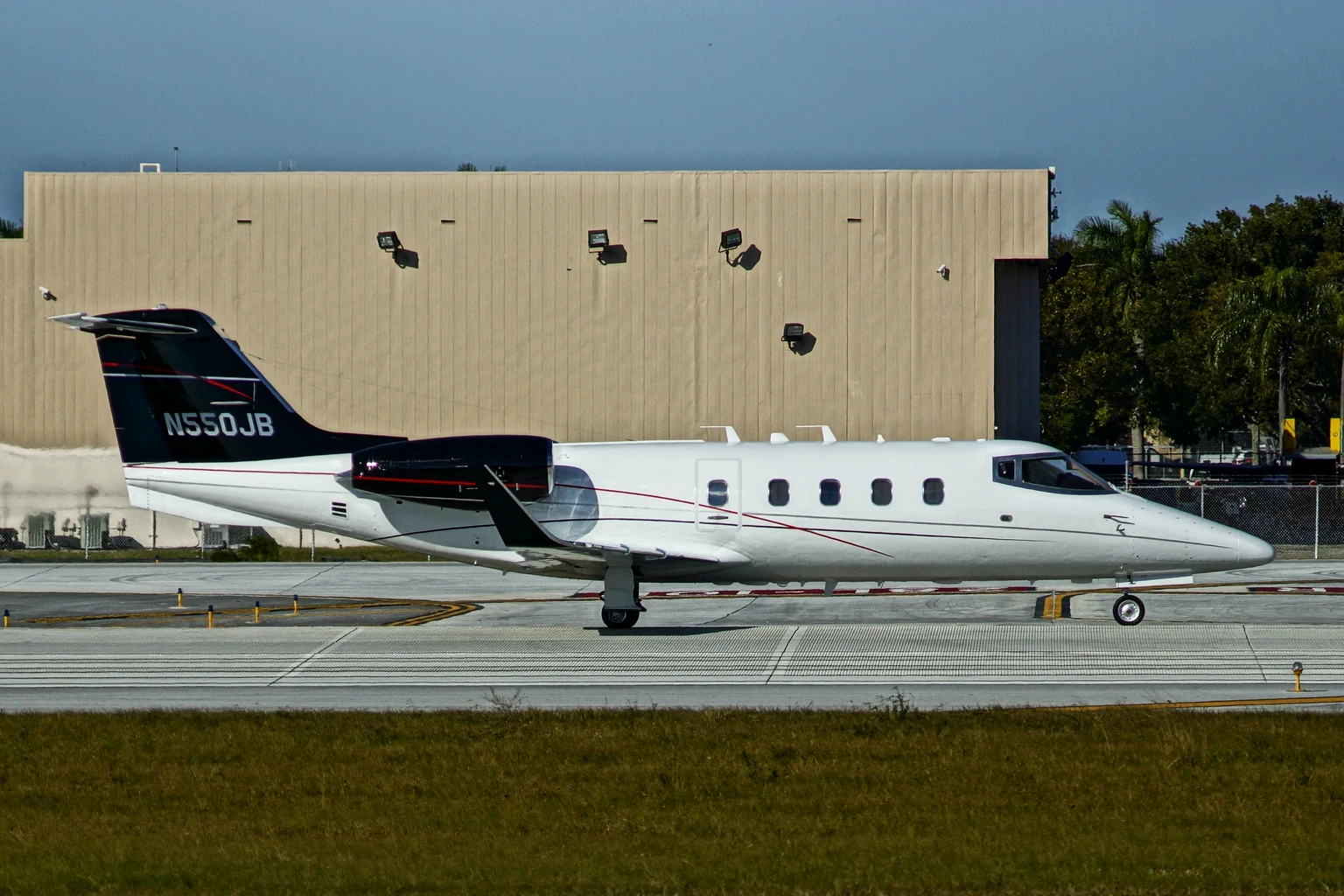 A white Learjet 55 with a black tail taxiing side-on in bright sun in front of a hangar.