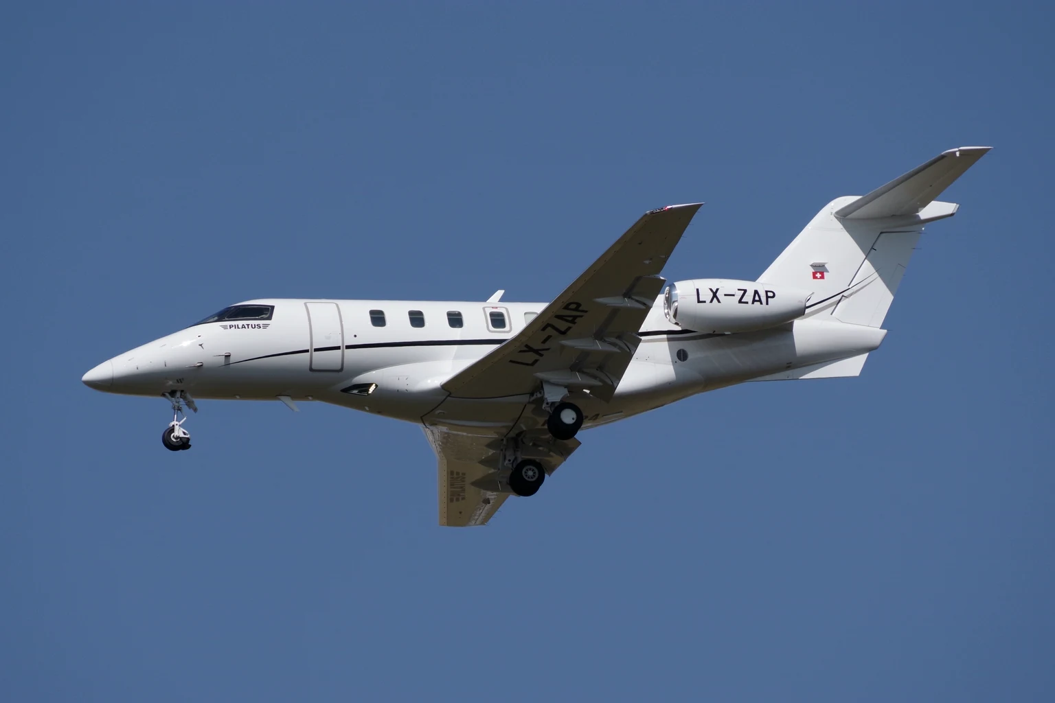 A white Pilatus PC-24 with a thin black cheatline on approach against a clear blue sky, seen from below with the gear down.