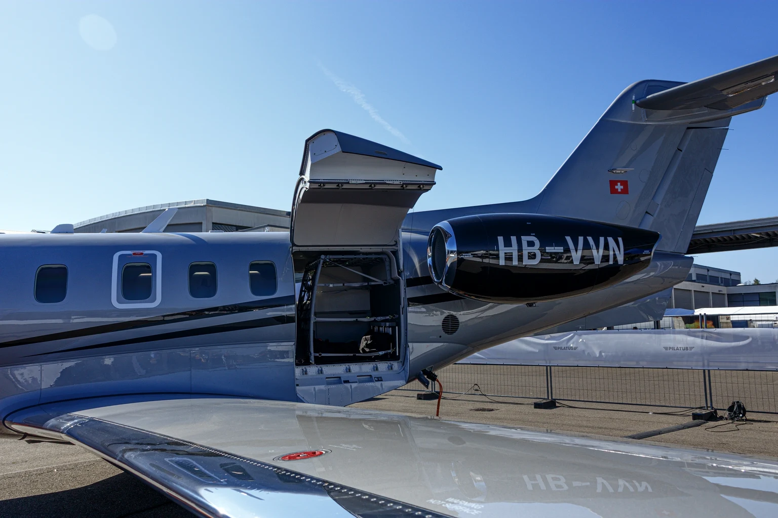 The rear of a silver PC-24 at a trade show with its large aft cargo door raised, showing the baggage room behind the cabin windows.