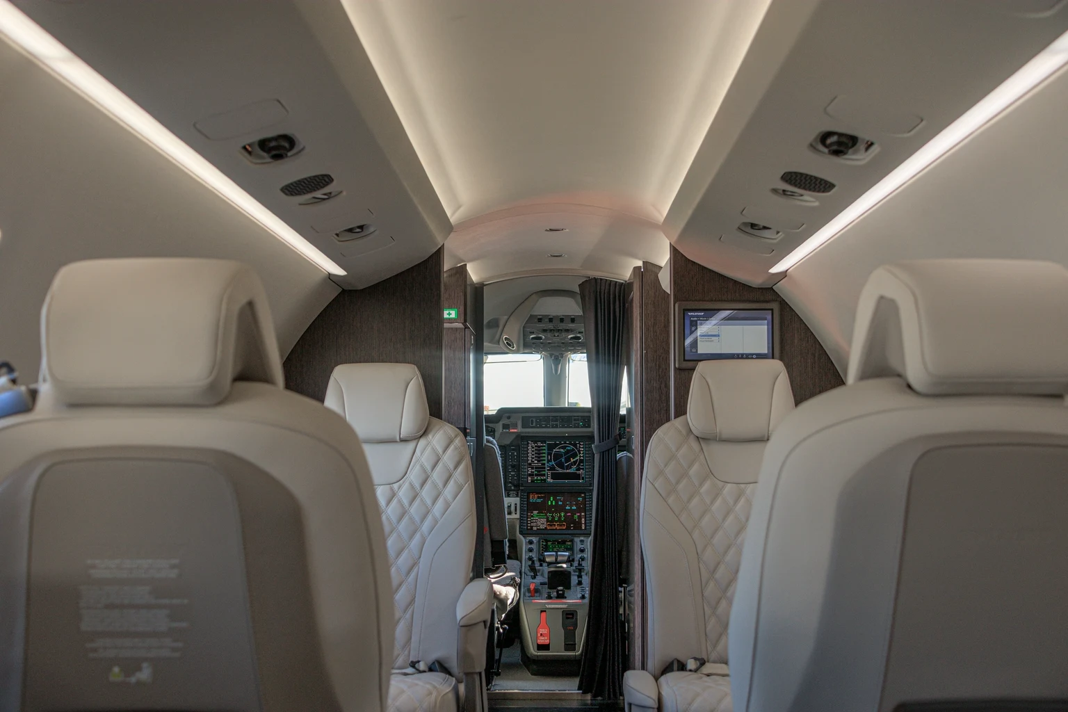 Looking forward through a PC-24 cabin: cream quilted leather seats, wood-grain bulkheads and the flight deck beyond a tied-back curtain.