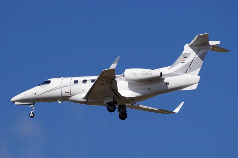 A plain white Embraer Phenom 300 on final approach against a deep blue sky, seen from below at a three-quarter angle with the gear down.