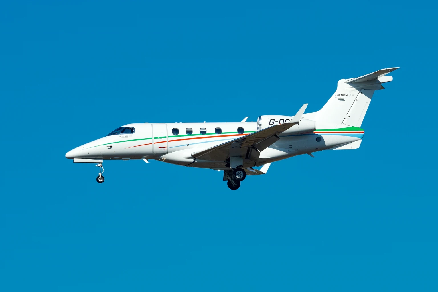 A white Phenom 300 with thin orange and green cheatlines on approach against a vivid blue sky, side-on.