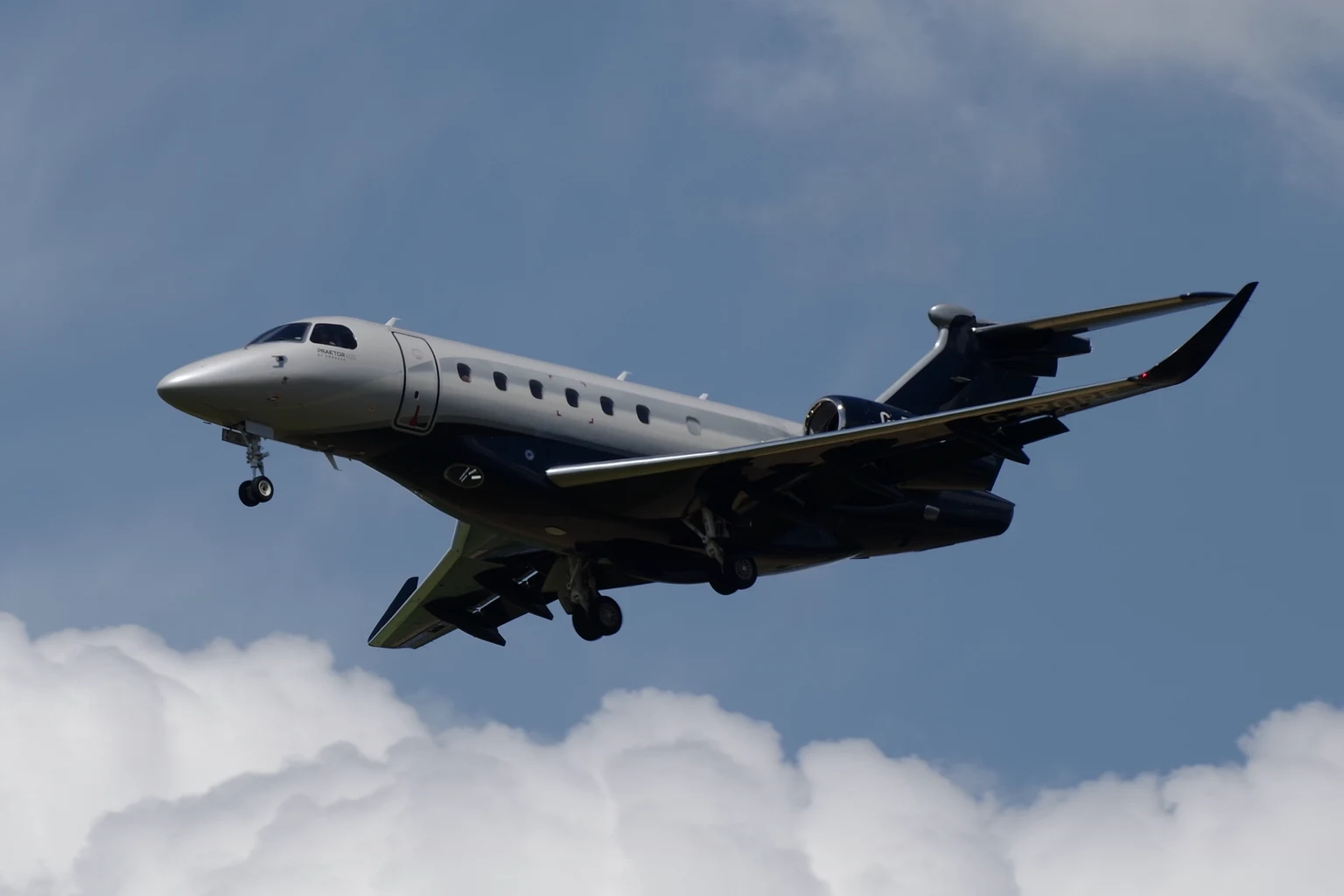 A silver Embraer Praetor 600 with a navy belly and tail on approach, blue sky above and a bank of white cumulus below.