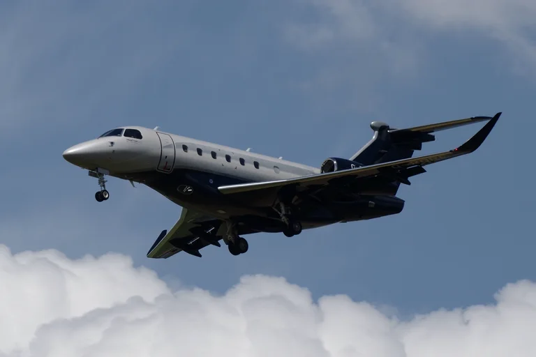 A silver Embraer Praetor 600 with a navy belly and tail on approach, blue sky above and a bank of white cumulus below.