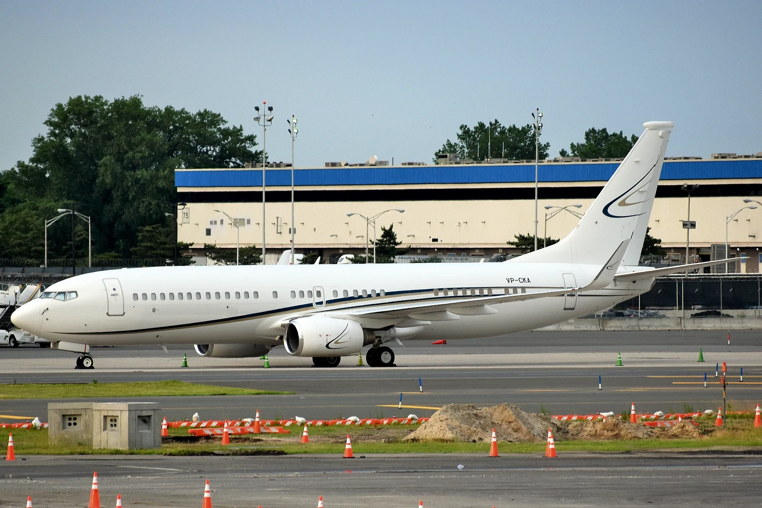 A white Boeing 737 business jet parked on a ramp at JFK, a hangar and trees behind it