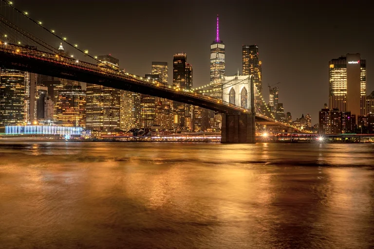 The Brooklyn Bridge and the Lower Manhattan skyline lit at night, reflected in the East River