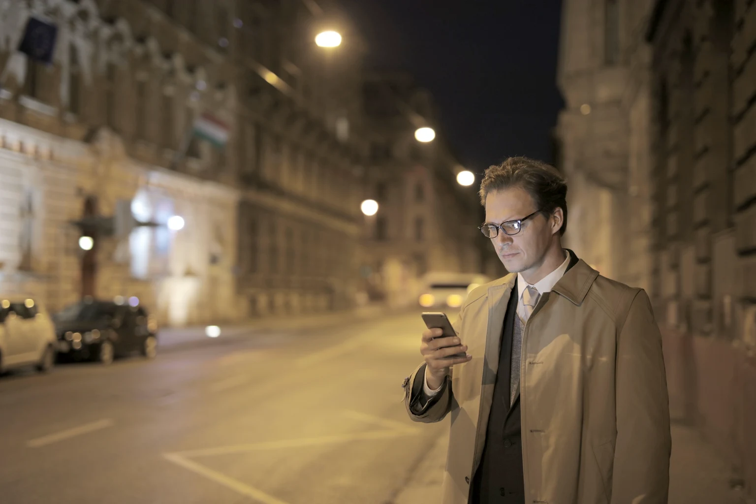 A man in a trench coat over a suit, reading his phone on a lamplit city street at night