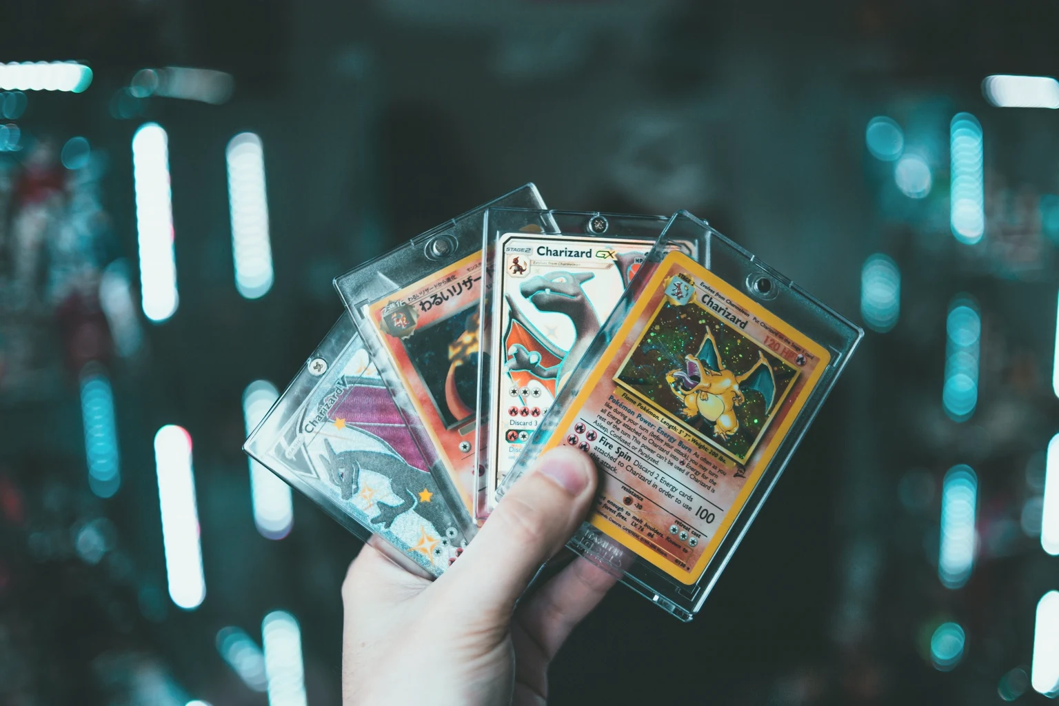 A hand holding four Charizard Pokémon cards fanned out in clear magnetic holders, against dark blurred lights