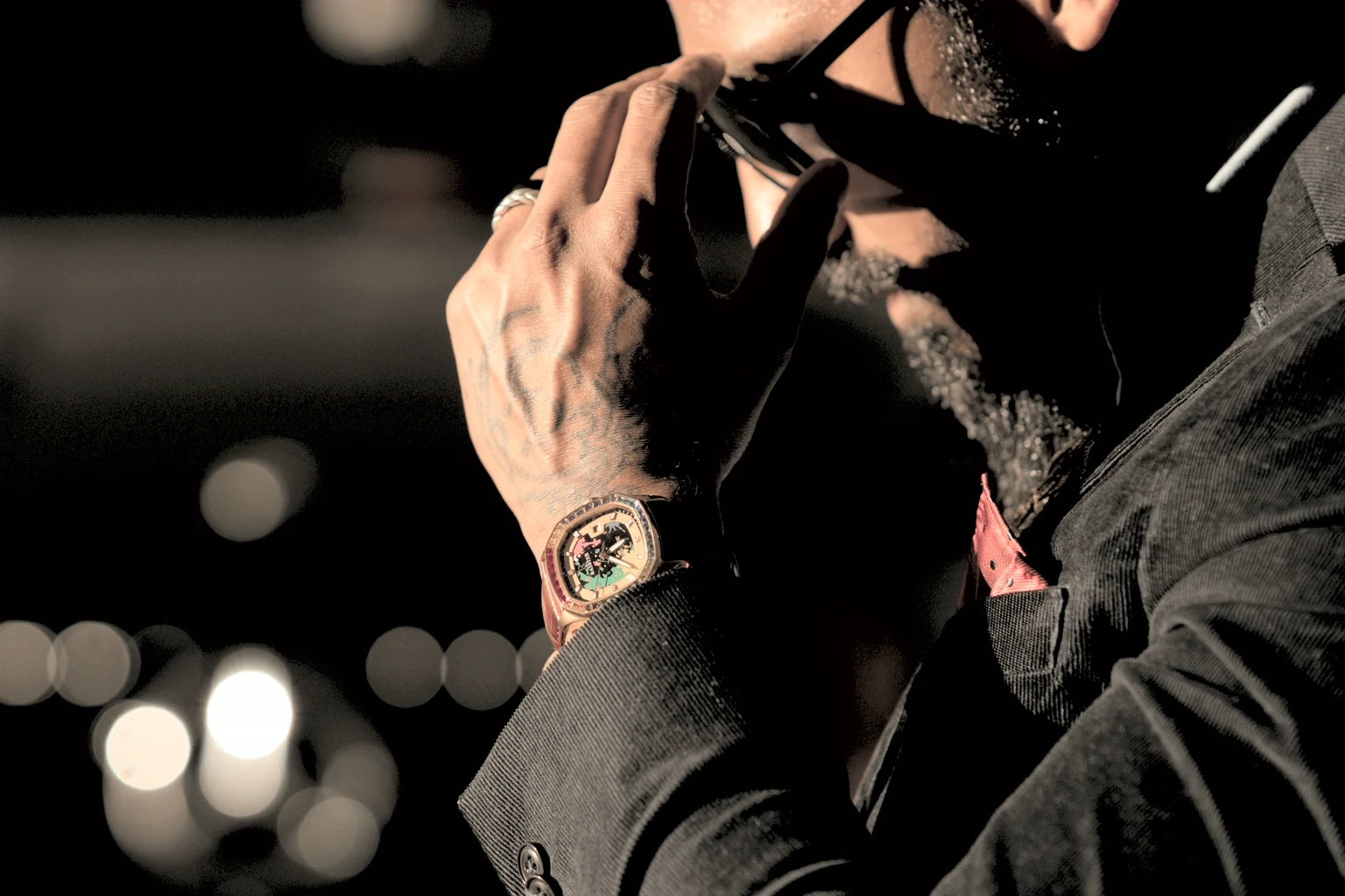A man in a dark jacket adjusting a gold wristwatch against city bokeh