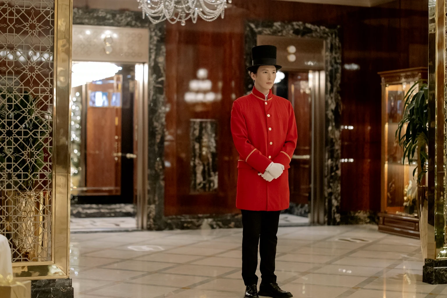 A hotel doorman in a red coat, top hat and white gloves waits in a marble lobby under a crystal chandelier