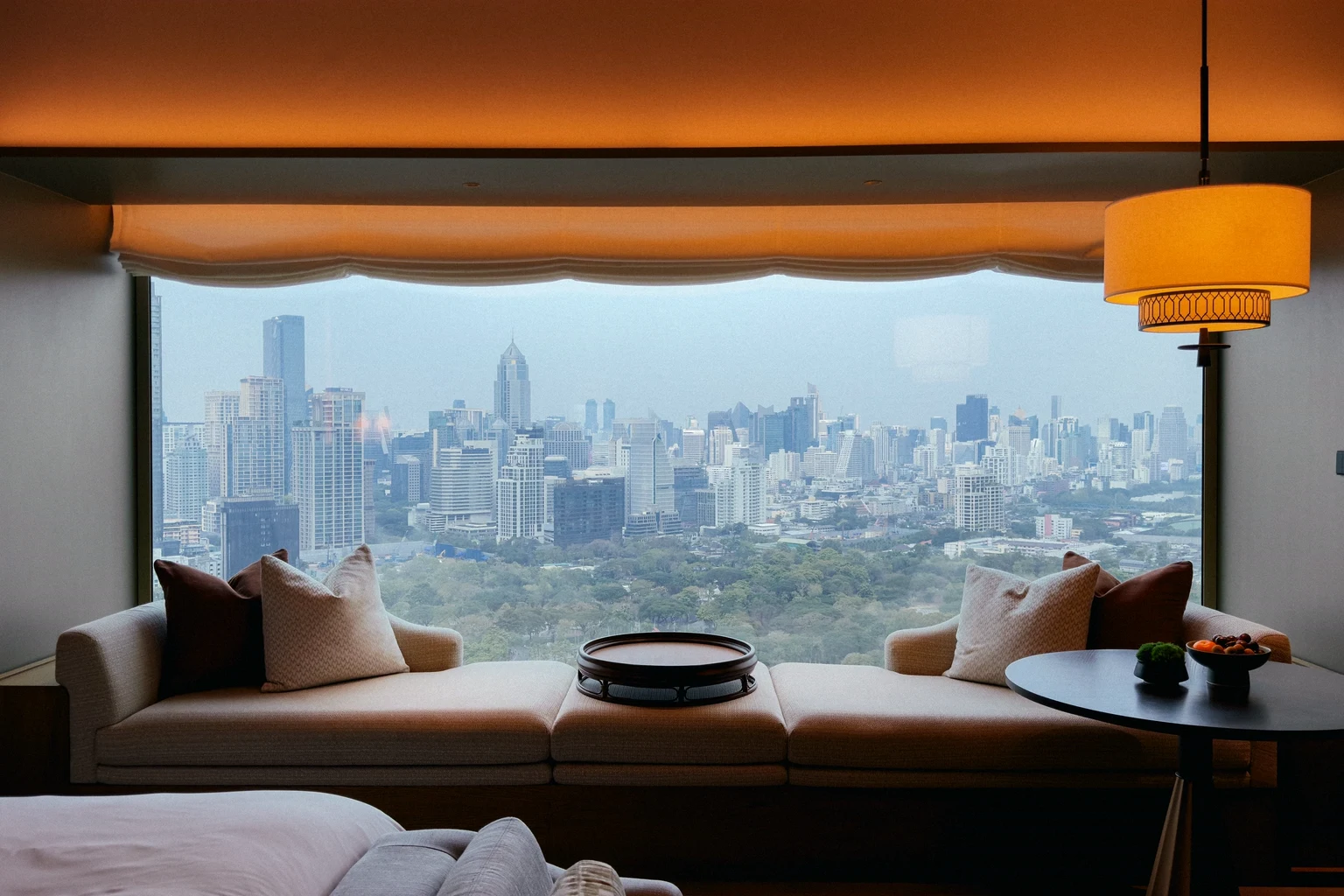 A hotel suite's window seat with cushions and a side table, looking out over a city skyline and a park