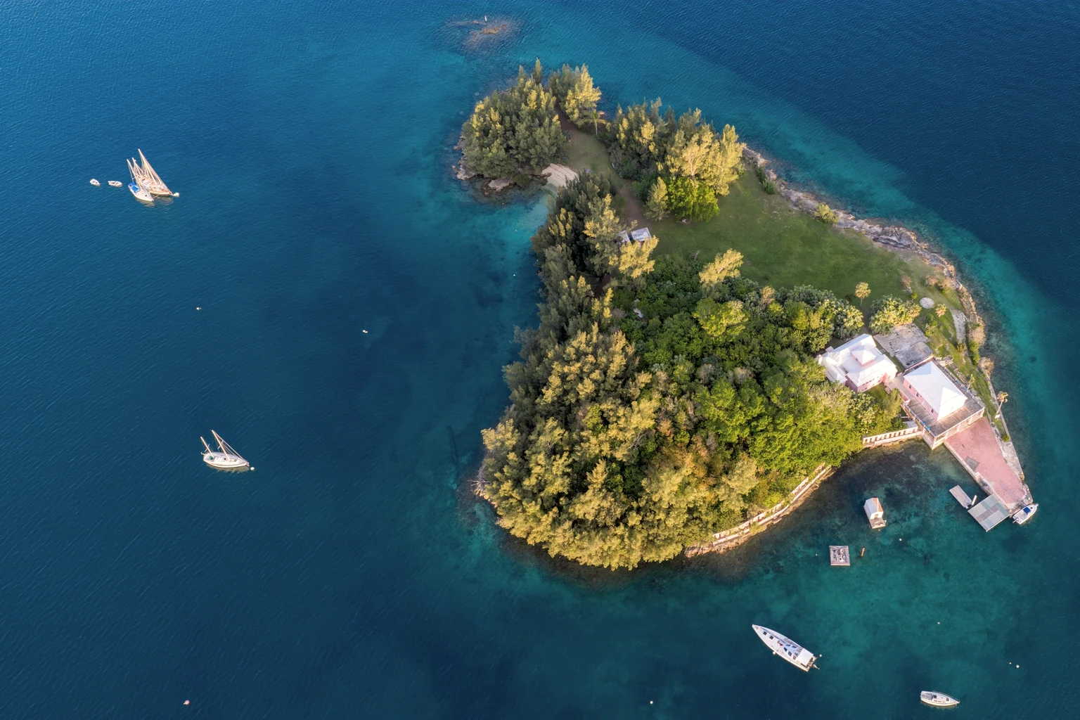 A small wooded private island with a house, a jetty and boats at anchor, seen from above in deep blue water