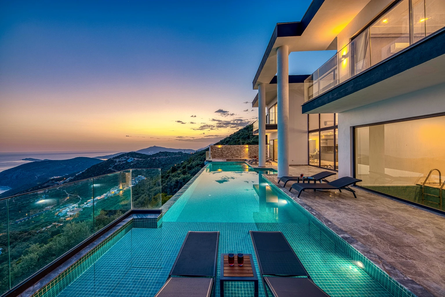 A modern villa with an infinity pool and two loungers on a hillside above the sea at dusk