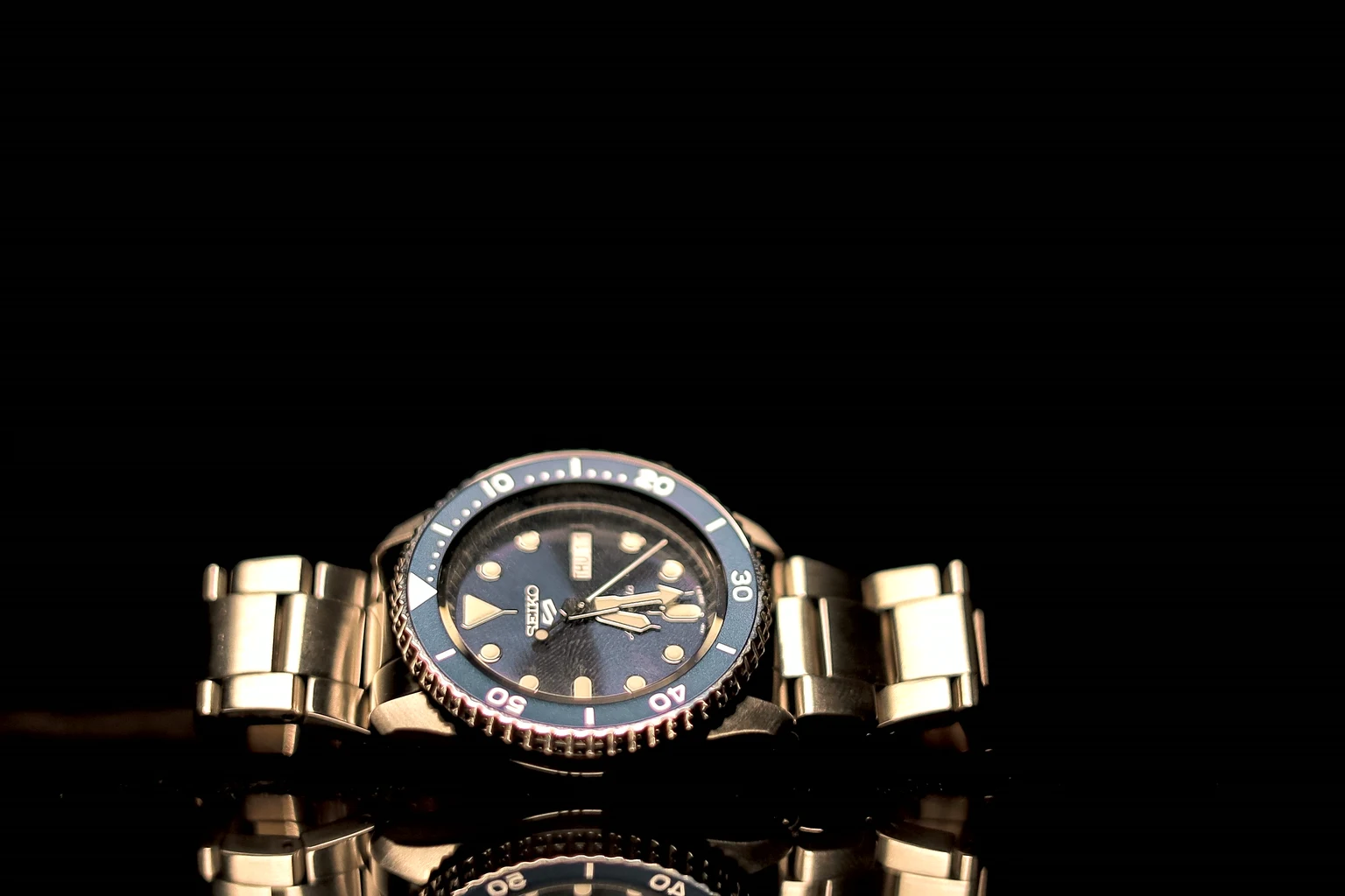 A gold watch with a black dial resting on a dark reflective surface