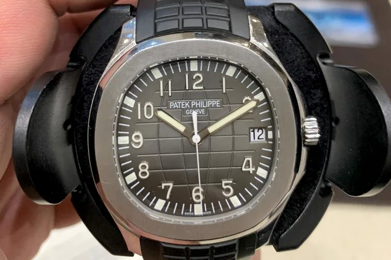 A steel Patek Philippe Aquanaut 5167A with a black embossed dial and black composite strap, held up in its travel case