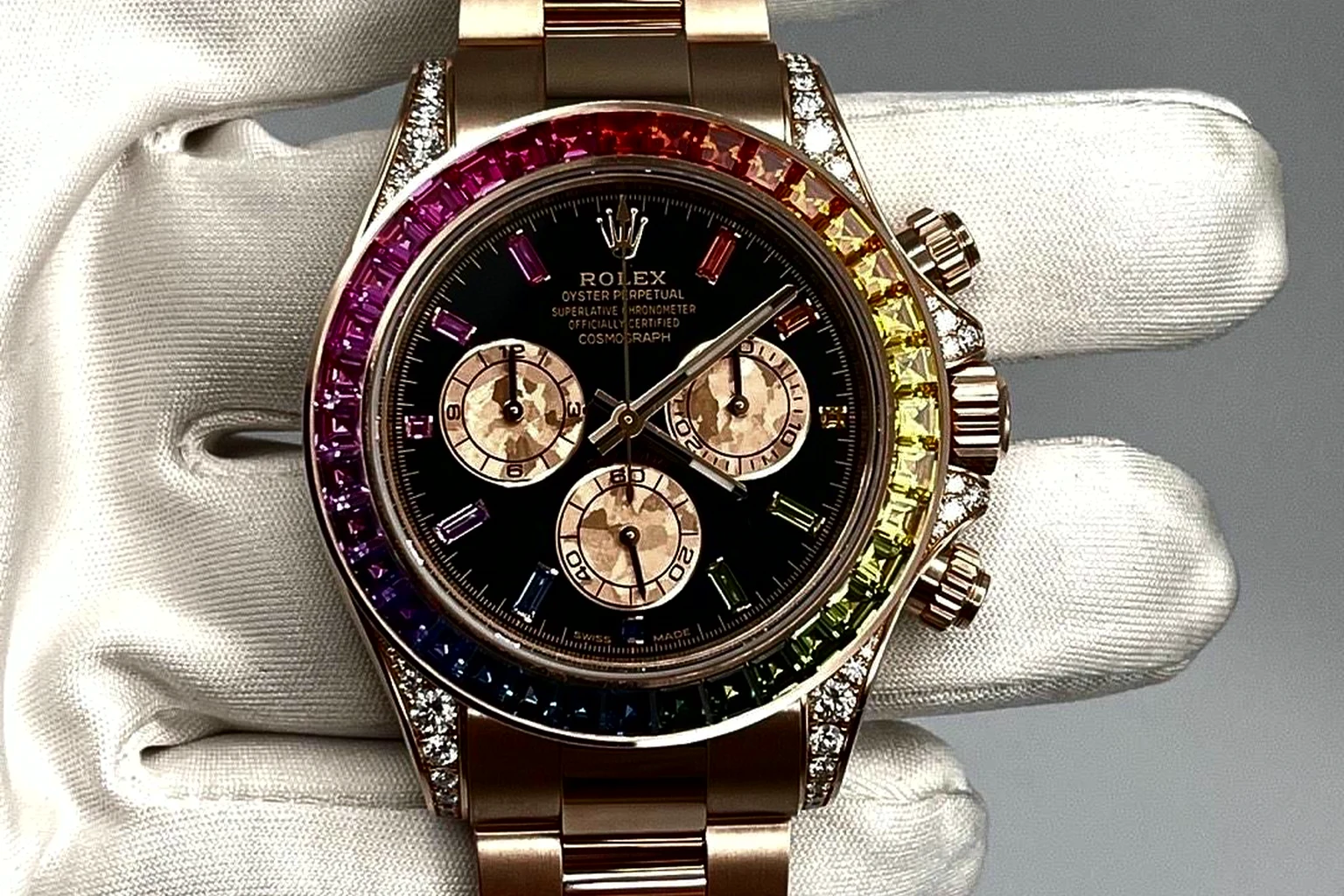 An Everose gold Rolex Cosmograph Daytona Rainbow, its bezel set with graduated coloured sapphires and a black dial, held in a white-gloved hand