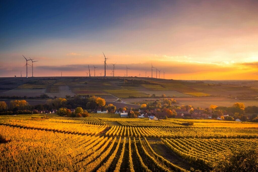 The Growing Trend: Why Ultra High Net Worth Individuals are Investing in Farmland and which billionaires have already invested? 1 Sunset over a rural landscape with wind turbines on a hill, patterned fields in the foreground, and a small village nestled among trees, reflecting the trend of Ultra High Net Worth individuals investing in far
