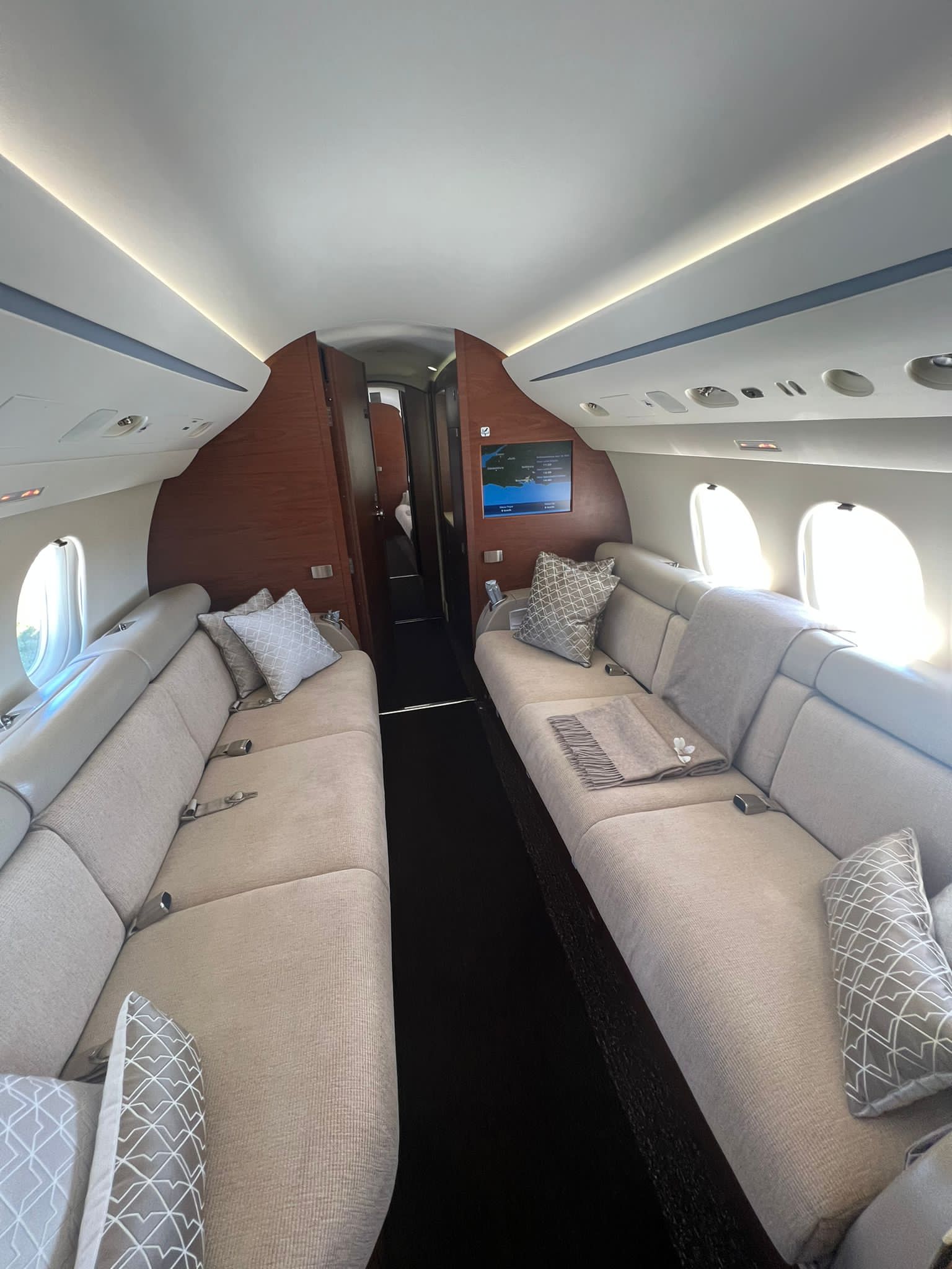 Interior of a Private Jet Athens, featuring beige leather seats, patterned cushions, elegant wood panelling, and windows on both sides. The aisle runs down the centre towards a closed door at the far end for added privacy.