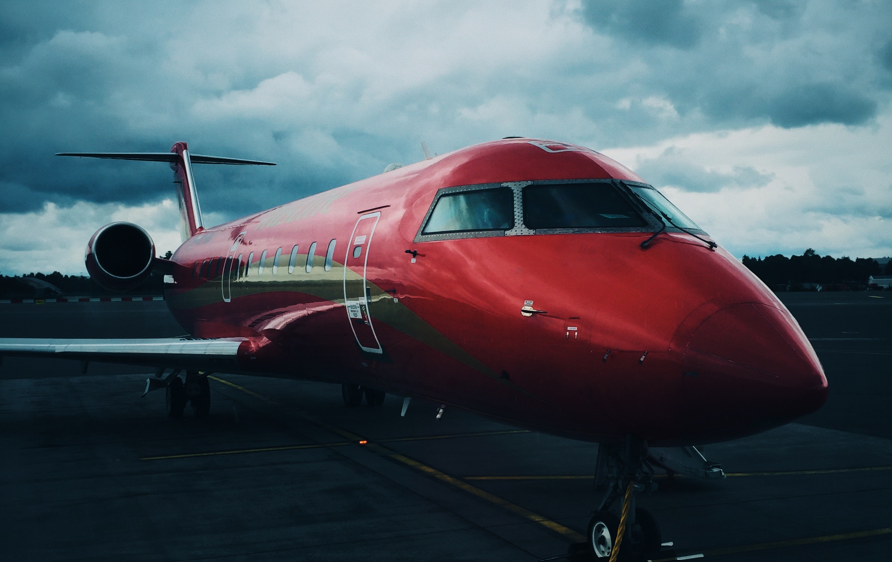 A red passenger aeroplane is parked on an airport tarmac under a cloudy sky, with trees and runway markings visible in the background.