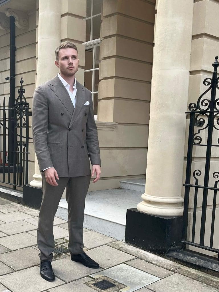 A man in a grey double-breasted suit with a white shirt and black shoes stands on a stone pavement in front of a beige building with tall pillars and black metal railings.