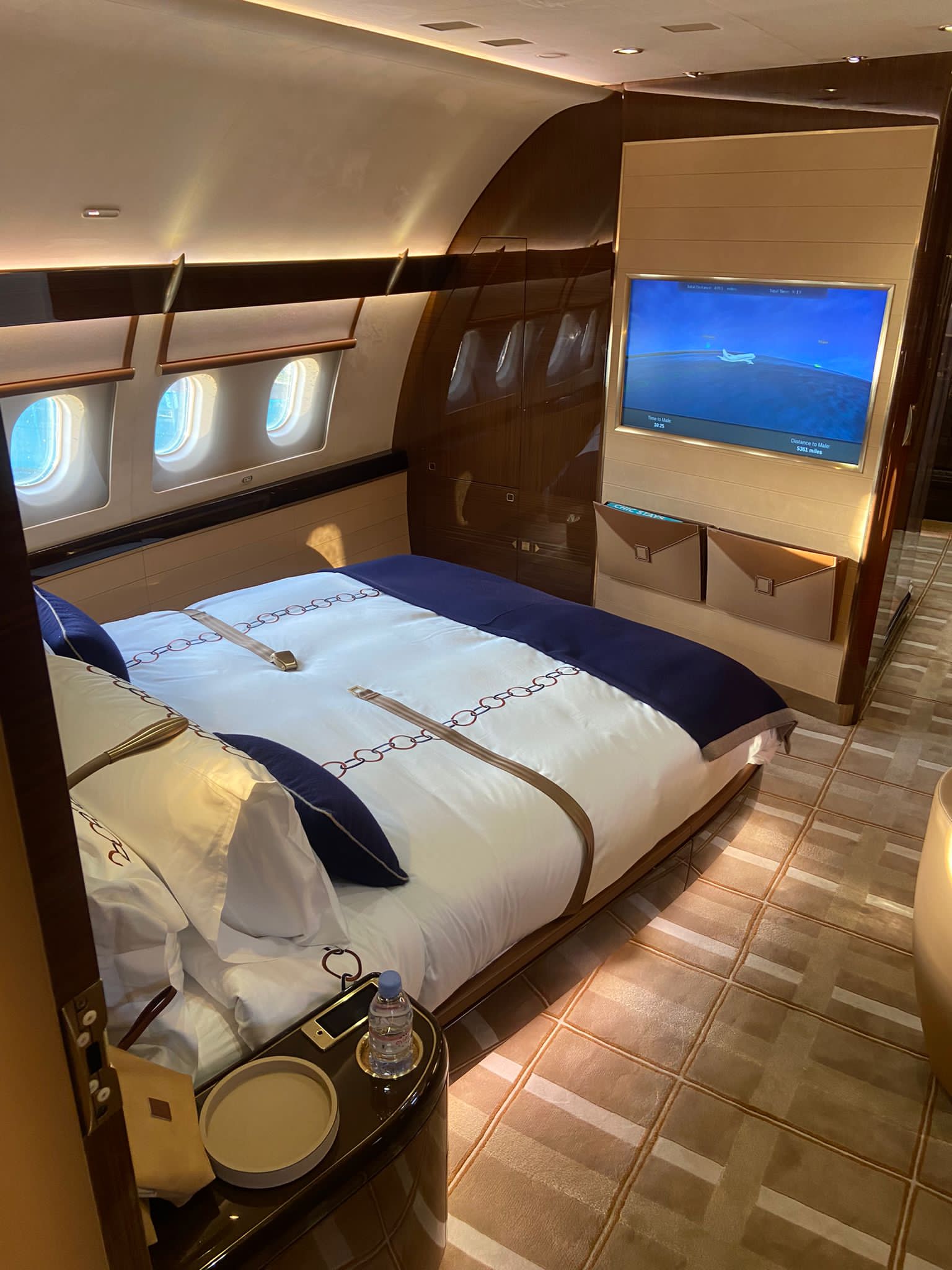 A luxurious private jet bedroom with a large bed, plush bedding, a small table with water, and a flat-screen telly displaying an aeroplane—perfect for relaxing on exclusive empty leg flights, all bathed in warm lighting with windows on one side.
